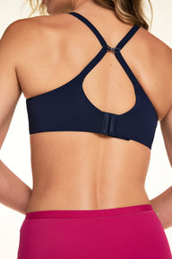 On a light studio backdrop, Alexis is shown from the back wearing the Base Unlined Bra in Navy with bright pink underwear. The image highlights the hook-and-eye closure, adjustable straps, and racerback conversion clip.