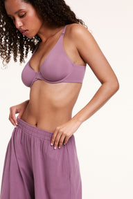 In a studio setting, Courtney wears the Base Unlined Bra in Mirage, shown from a side angle highlighting the unlined underwire cups, smooth microfiber fabric, and supportive band. The front-adjusting strap is visible at the shoulder.