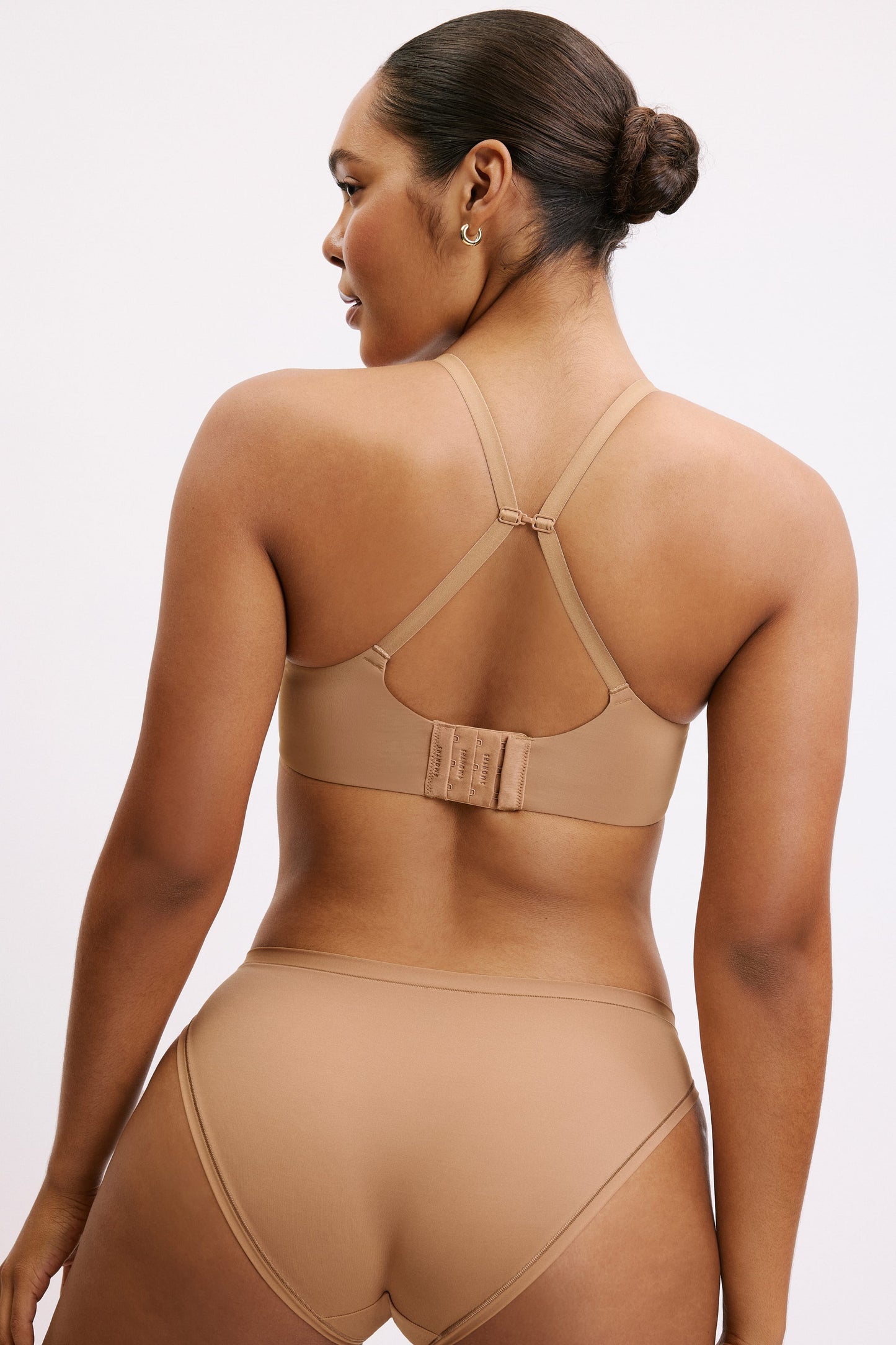 On a light studio backdrop, Amber wears the Base Unlined Bra in Tan, shown from the back with the J-hook fastened into a racerback for added support.