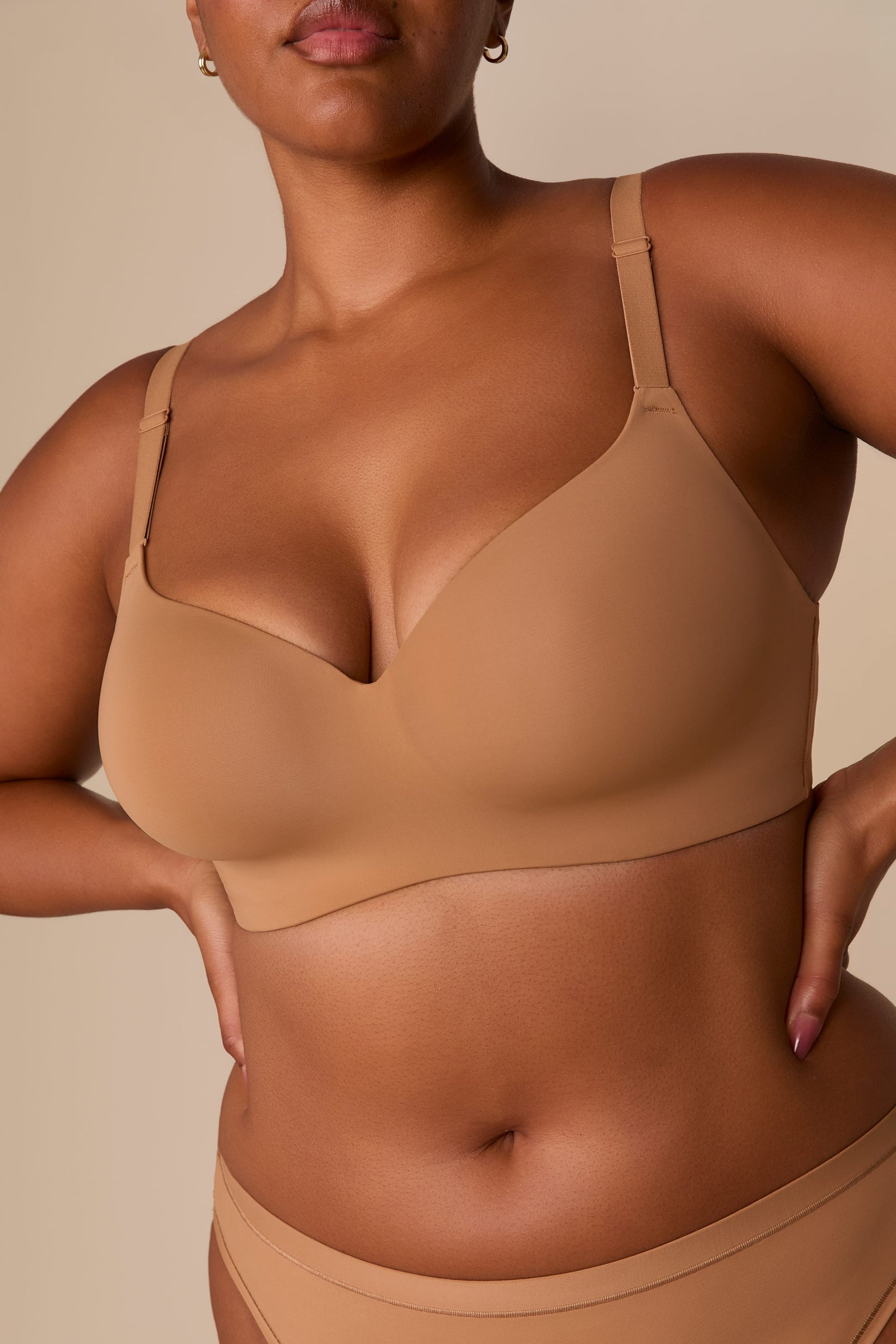On a neutral beige backdrop, a close-up of Zarria wearing the Base Wirefree T-Shirt Bra in Tan showcases the smooth molded cups, wireless construction, and adjustable straps.