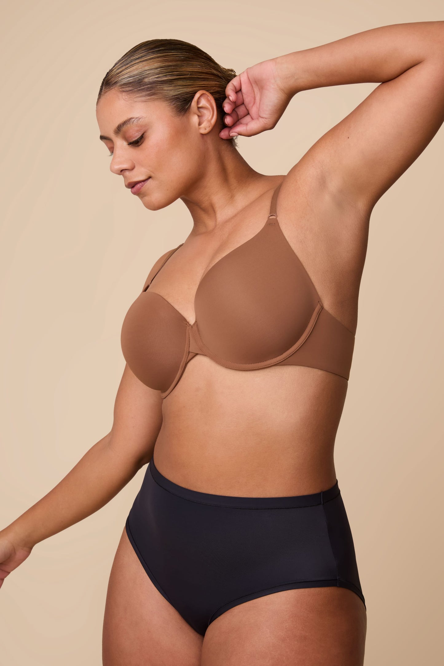 On a neutral beige backdrop, India wears the Base T-Shirt Bra in Brown, shown in profile to highlight the lightly lined cups, underwire support, and smooth microfiber fabric.