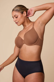 On a neutral beige backdrop, India wears the Base T-Shirt Bra in Brown, shown in profile to highlight the lightly lined cups, underwire support, and smooth microfiber fabric.