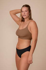 On a neutral beige backdrop, Sonnie wears the Bliss Bikini in Black with a matching bralette in a warm brown shade, standing in a three-quarter side pose that highlights the smooth waistband and comfortable fit.