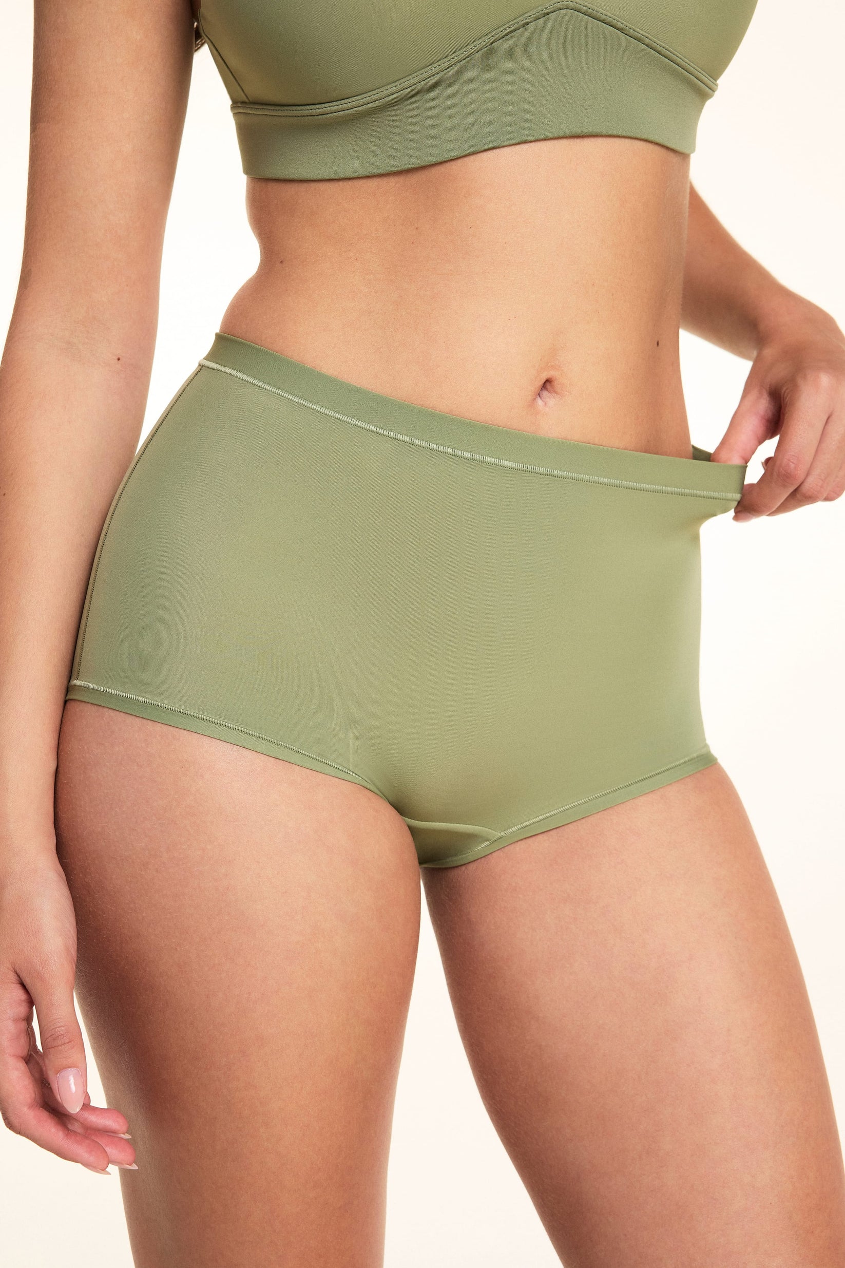 On a white studio backdrop, Laura wears the Bliss Boyshort in Dill and gently stretches the waistband to show the soft, flexible microfiber fabric and high-rise fit.