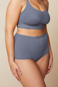 On a neutral beige backdrop, a close-up of Summerlyn wearing the Bliss Boyshort in Steel showcases the high-rise fit and smooth fabric at the waist and leg openings.