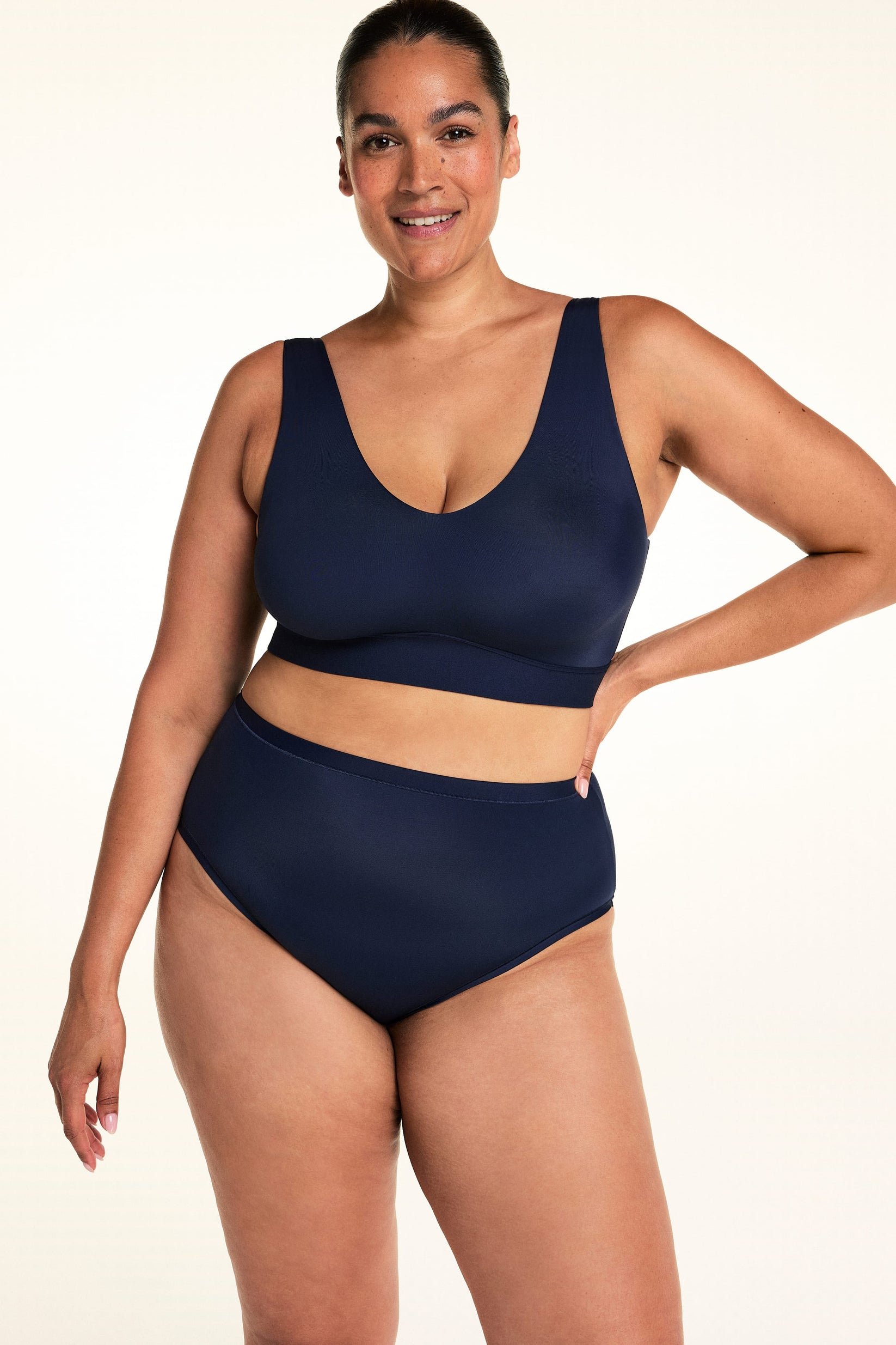 On a light studio backdrop, Jennifer wears the Bliss Bralette in Navy with matching underwear. Standing with one hand at her waist, she shows the V-neckline, wireless support, and smooth silhouette of the bralette.