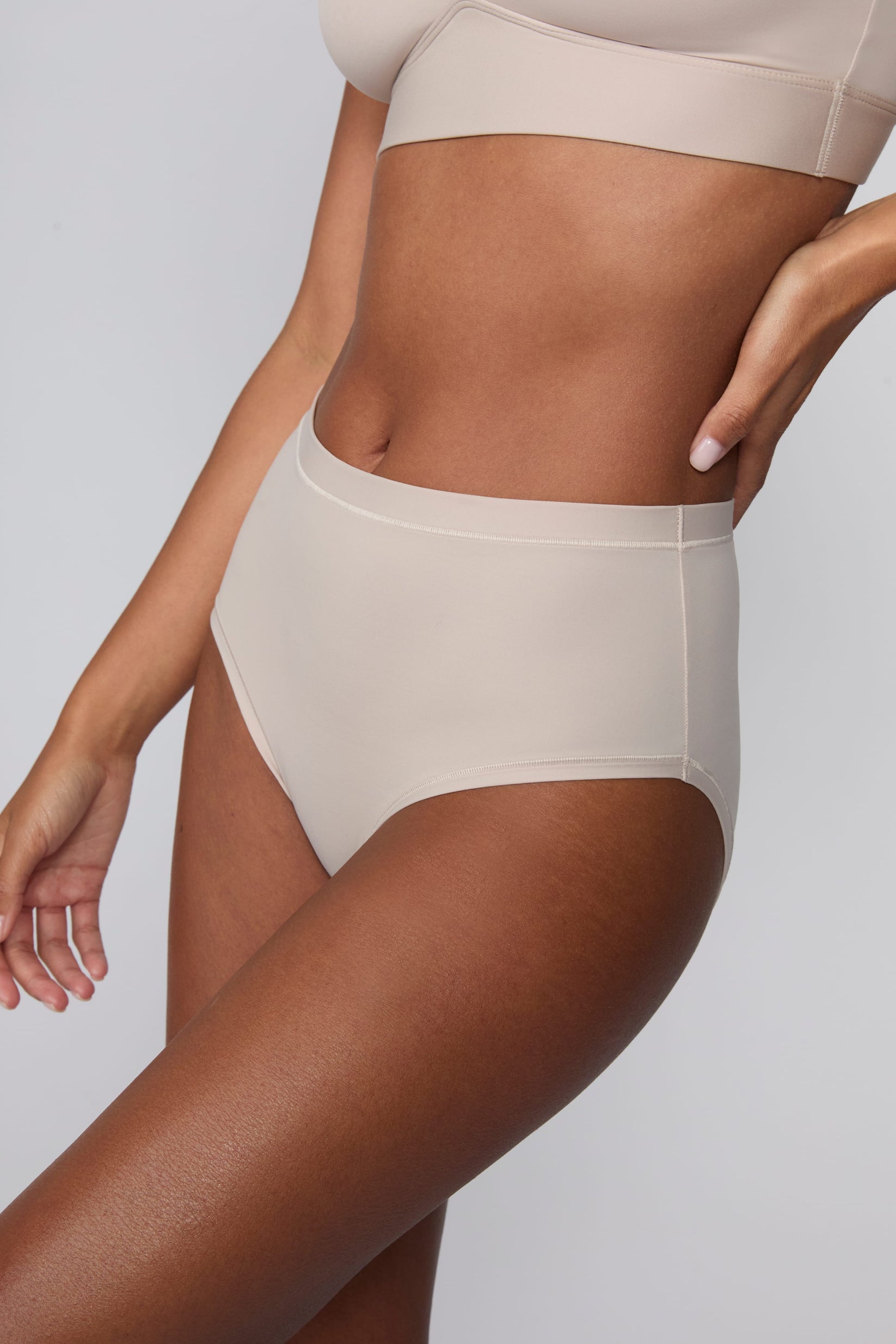 In a studio setting, Courtney wears the Bliss Highwaist Brief in Pebble. A side-angle close-up highlights the smooth fabric, high-rise waistband, and full-coverage design.