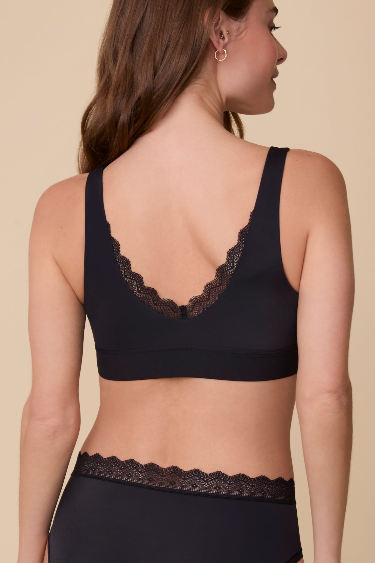 On a neutral beige backdrop, Gracie wears the Bliss Lace Bralette in Black with coordinating underwear, showing the back view with a low scoop back, scalloped lace detail, and smooth supportive band.