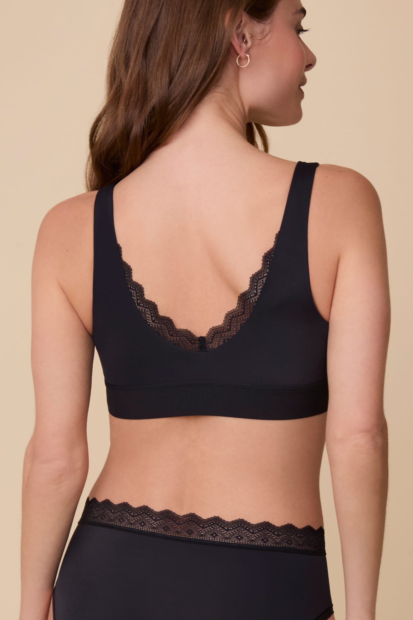 On a neutral beige backdrop, Gracie wears the Bliss Lace Bralette in Black with coordinating underwear, showing the back view with a low scoop back, scalloped lace detail, and smooth supportive band.