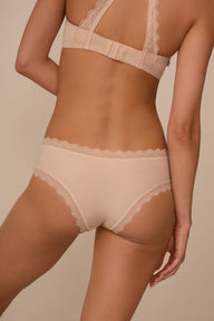 On a neutral studio backdrop, Marcela wears the Bliss Lace Cheeky in Beige with a matching beige bra. The back view highlights cheeky back coverage, smooth fabric, and scalloped lace trim along the waistband and leg openings.