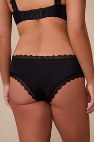 On a neutral studio backdrop, Sonnie wears the Bliss Lace Cheeky in Black. The close-up back view highlights the smooth fabric, cheeky coverage, and scalloped lace trim along the waistband and leg openings.
