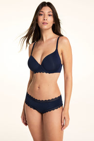 On a light studio backdrop, Laura wears the Bliss Lace Cheeky in Navy with a matching navy bra. The front view highlights the smooth fabric and scalloped lace trim along the waistband and leg openings.