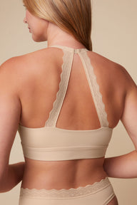 On a neutral beige backdrop, a back view of Megan wearing the Bliss Lace Halterneck Bralette in Beige showcases the lace halterneck straps, smooth back band, and lace-trimmed matching underwear.