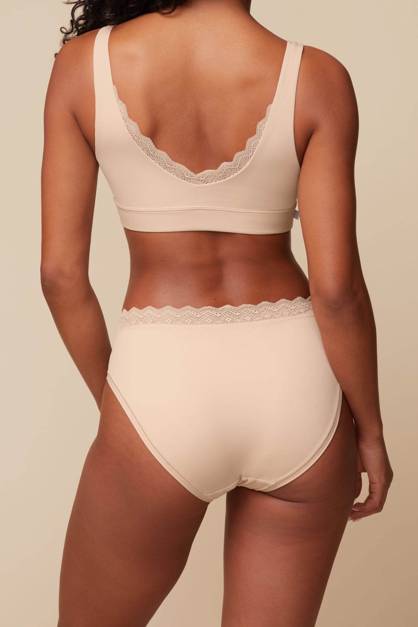 On a neutral beige backdrop, a back view of Ally wearing the Bliss Lace Highwaist Brief in Beige showcases the full-coverage back and lace-trim waistband paired with the matching Bliss Lace Bralette.