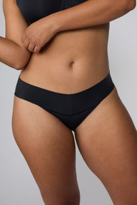 On a light gray studio backdrop, India wears the Bliss Low Rise Bikini in Black. A close-up front view highlights the low-rise waistband, smooth construction, and bikini silhouette.