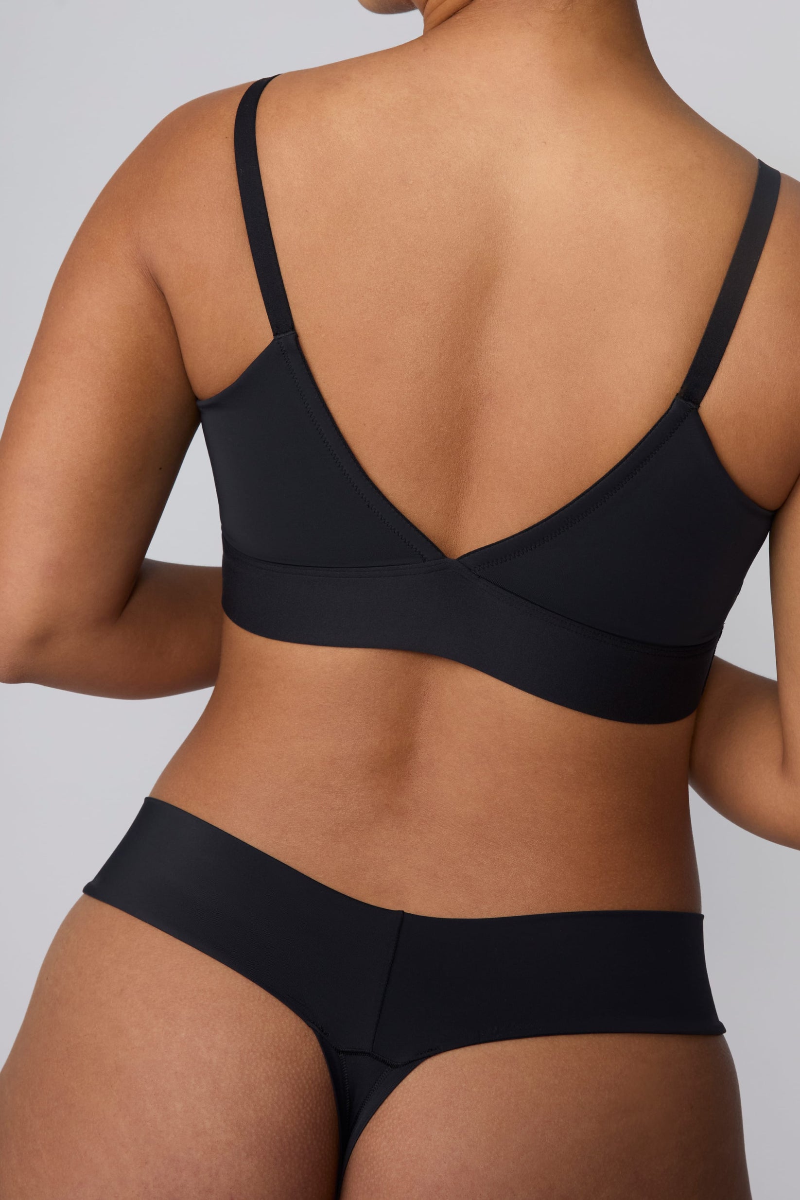On a light gray studio backdrop, India wears the Bliss Low Rise Thong in Black with the matching Bliss Bralette in Black. The back view highlights the thong silhouette, smooth waistband, and minimal back coverage.