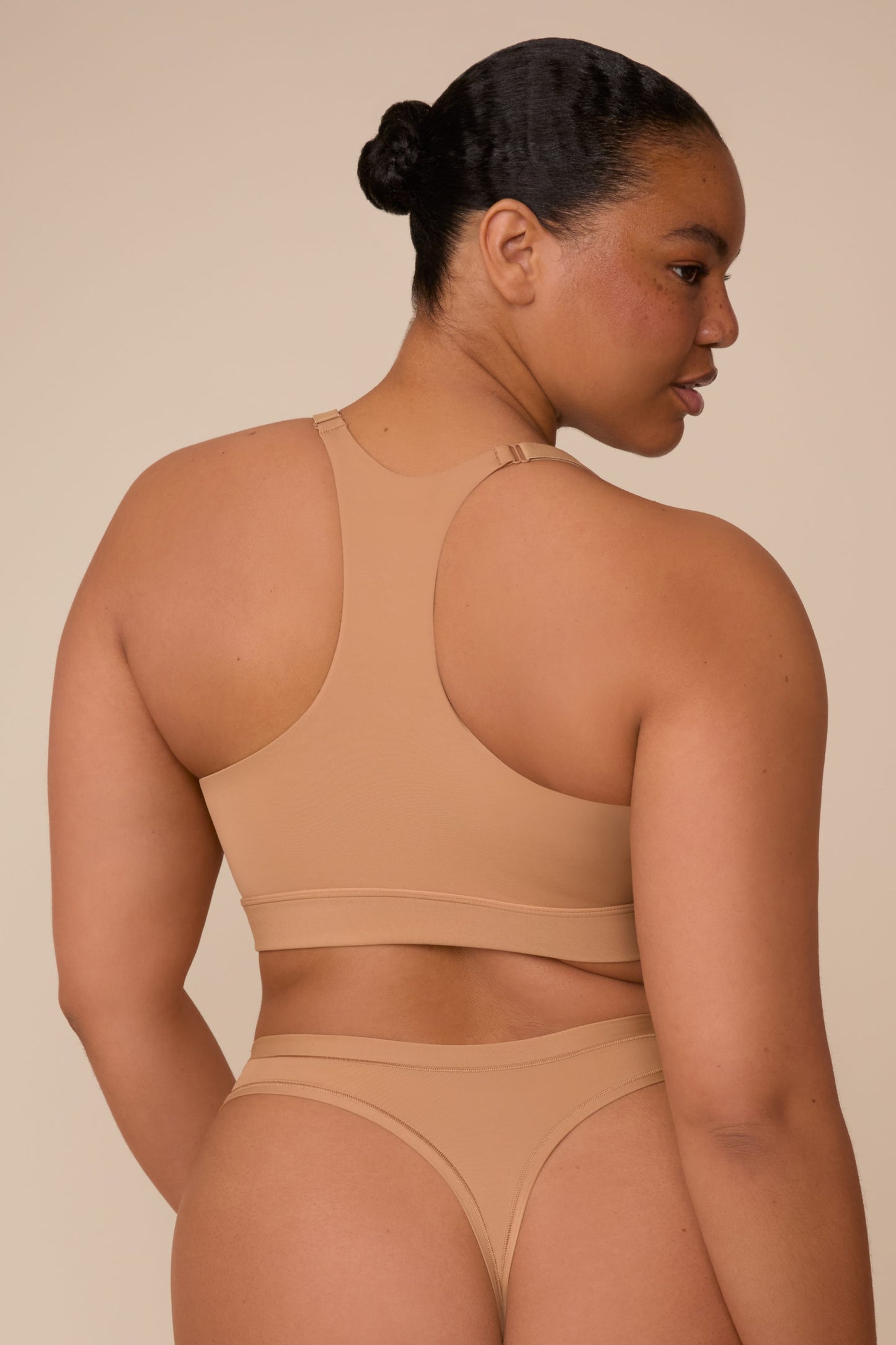 On a neutral beige backdrop, a back view of Zarria wearing the Bliss Racerback Bralette in Tan highlights the racerback design, adjustable straps, and wide underband.