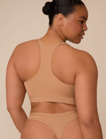 On a neutral beige backdrop, a back view of Zarria wearing the Bliss Racerback Bralette in Tan highlights the racerback design, adjustable straps, and wide underband. sizing: Zarria wears size XL