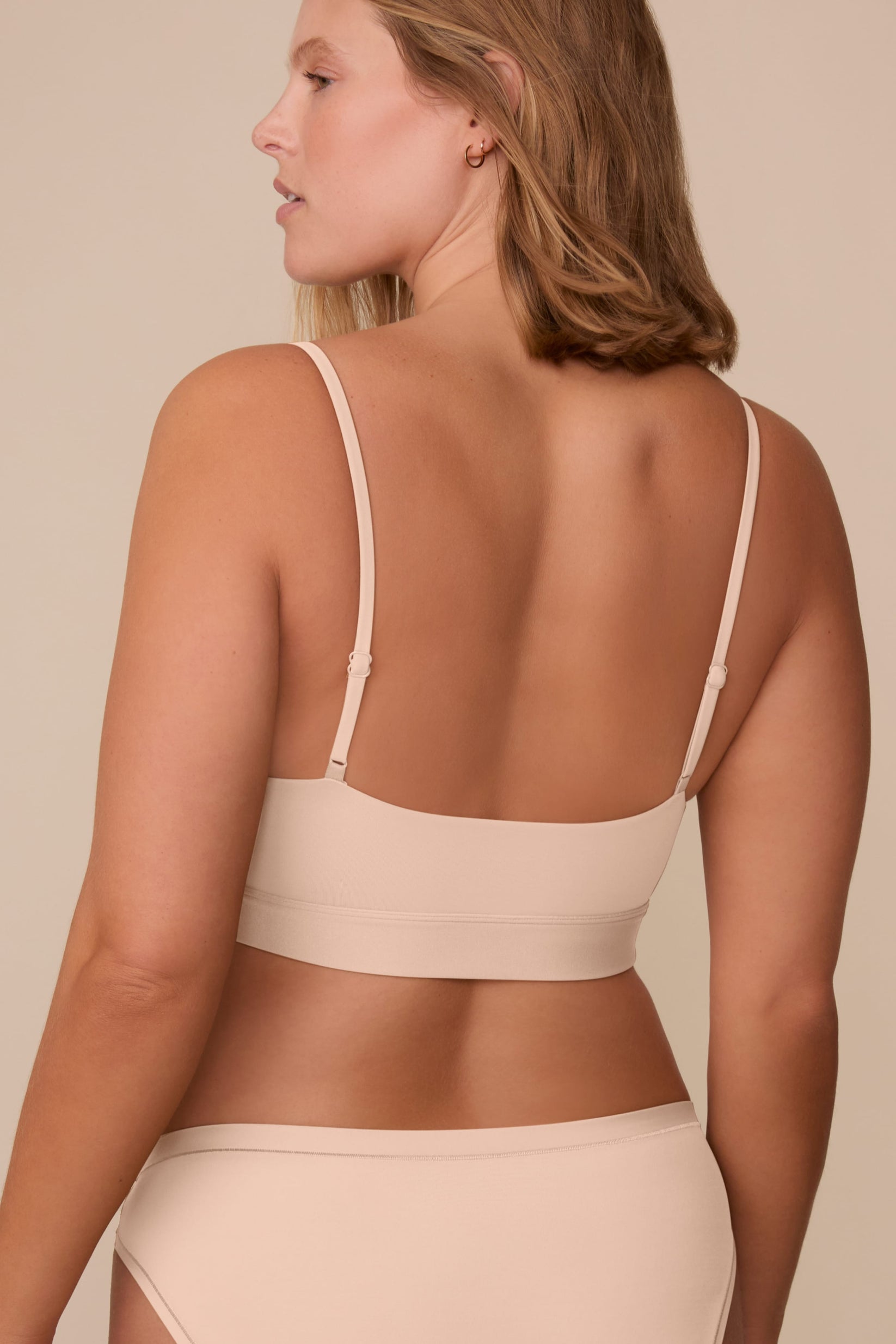 On a neutral beige backdrop, a back view of Sonnie wearing the Bliss Scoop Adjustable Bralette in Beige highlights the adjustable straps, wide underband, and smooth back design.
