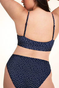 On a light studio backdrop, Alessandra is shown from the back wearing the Bliss Scoop Bralette in Navy Dot with matching Navy Dot underwear. The image highlights the scoop back, adjustable straps, wide underband, and allover white dot print.