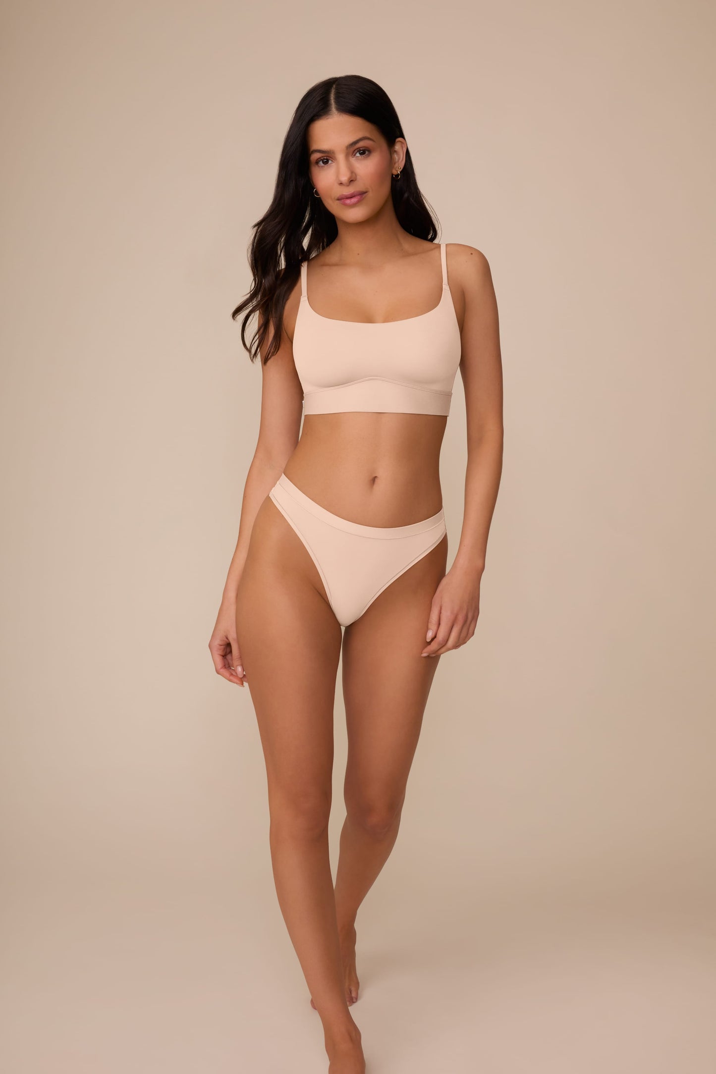 On a neutral beige backdrop, Marcela wears the Bliss Thong in Beige with a matching Bliss bralette, showing the low-rise silhouette, smooth fabric, and comfortable fit in a full-body view.