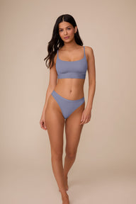 On a neutral beige backdrop, Marcela wears the Bliss Thong in Steel with a matching Bliss bralette, walking toward the camera and showcasing the low-rise fit, smooth waistband, and lightweight feel.