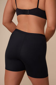 On a neutral studio backdrop, Sonnie wears the Bliss Under Short in Black. The back view highlights the smooth waistband, full back coverage, and thigh-length design.