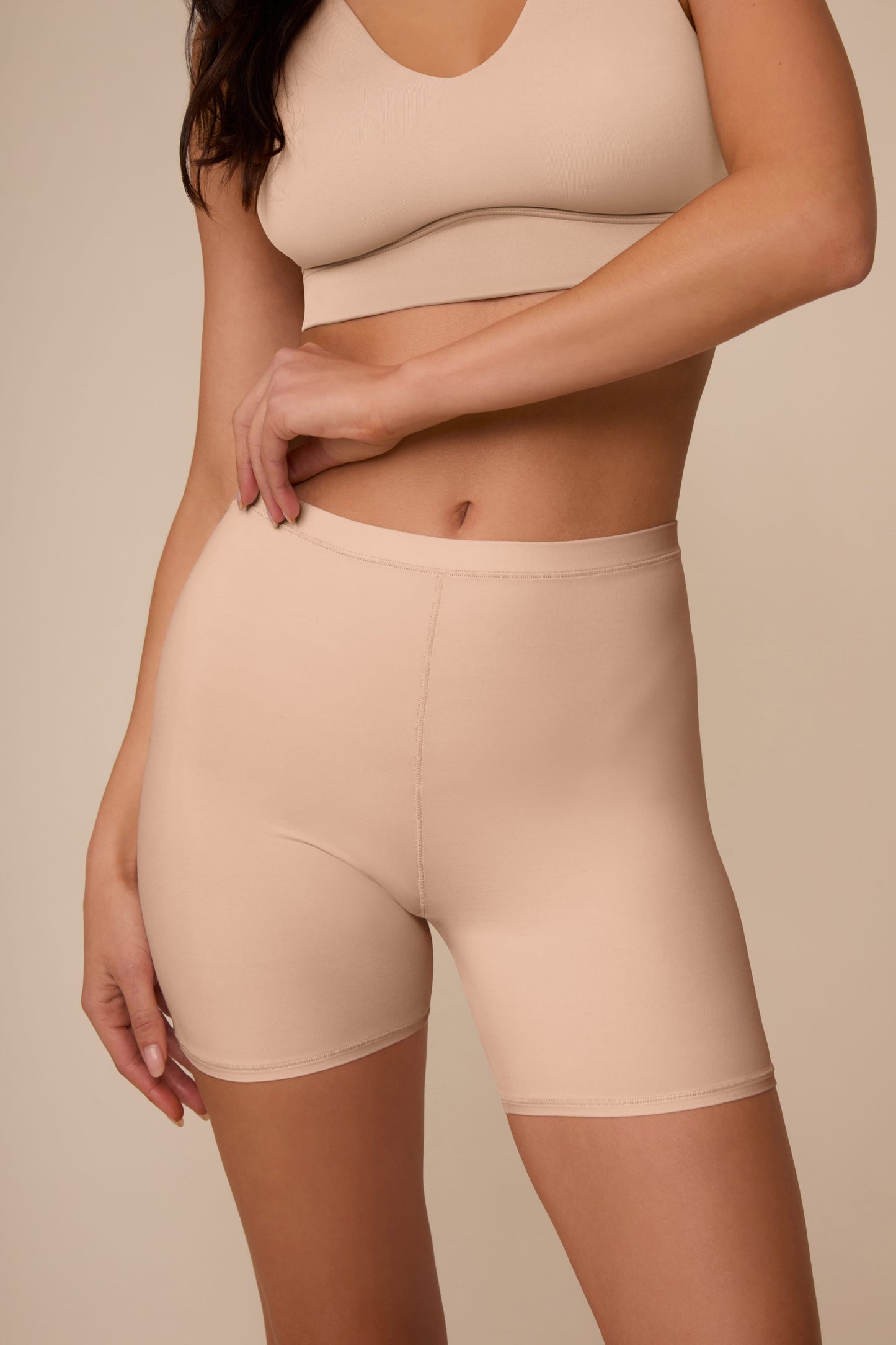 On a neutral studio backdrop, Marcela wears the Bliss Under Short in Beige with a matching beige bralette. The close-up front view highlights the smooth waistband, soft fabric, and streamlined fit through the hips and thighs.