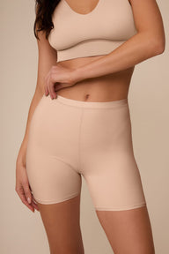 On a neutral studio backdrop, Marcela wears the Bliss Under Short in Beige with a matching beige bralette. The close-up front view highlights the smooth waistband, soft fabric, and streamlined fit through the hips and thighs.
