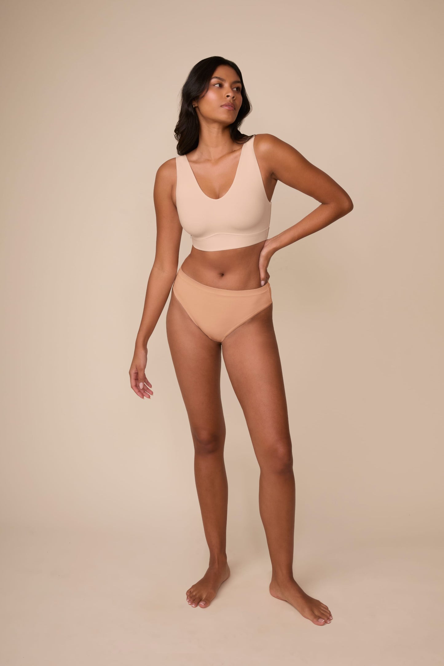 On a neutral beige backdrop, Victoria wears the Cloud Cotton Highwaist Thong in Dune with a coordinating bralette, showcasing the high-rise silhouette and thong back coverage in a full-body profile pose.