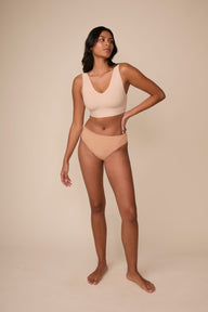 On a neutral beige backdrop, Victoria wears the Cloud Cotton Highwaist Thong in Dune with a coordinating bralette, showcasing the high-rise silhouette and thong back coverage in a full-body profile pose.