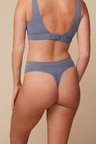 On a neutral beige backdrop, a back view of Megan wearing the Cloud Cotton Highwaist Thong in Slate showcases the thong back silhouette, soft cotton fabric, and smooth waistband. The matching Cloud Cotton Bra features a scoop back and hook-and-eye closure.