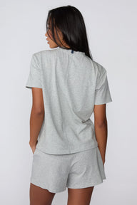 On a light gray studio backdrop, Amber wears the Cloud Cotton Pajama Shirt in Heather Grey with matching pajama shorts. The back view highlights the relaxed silhouette, short sleeves, and soft cotton fabric.