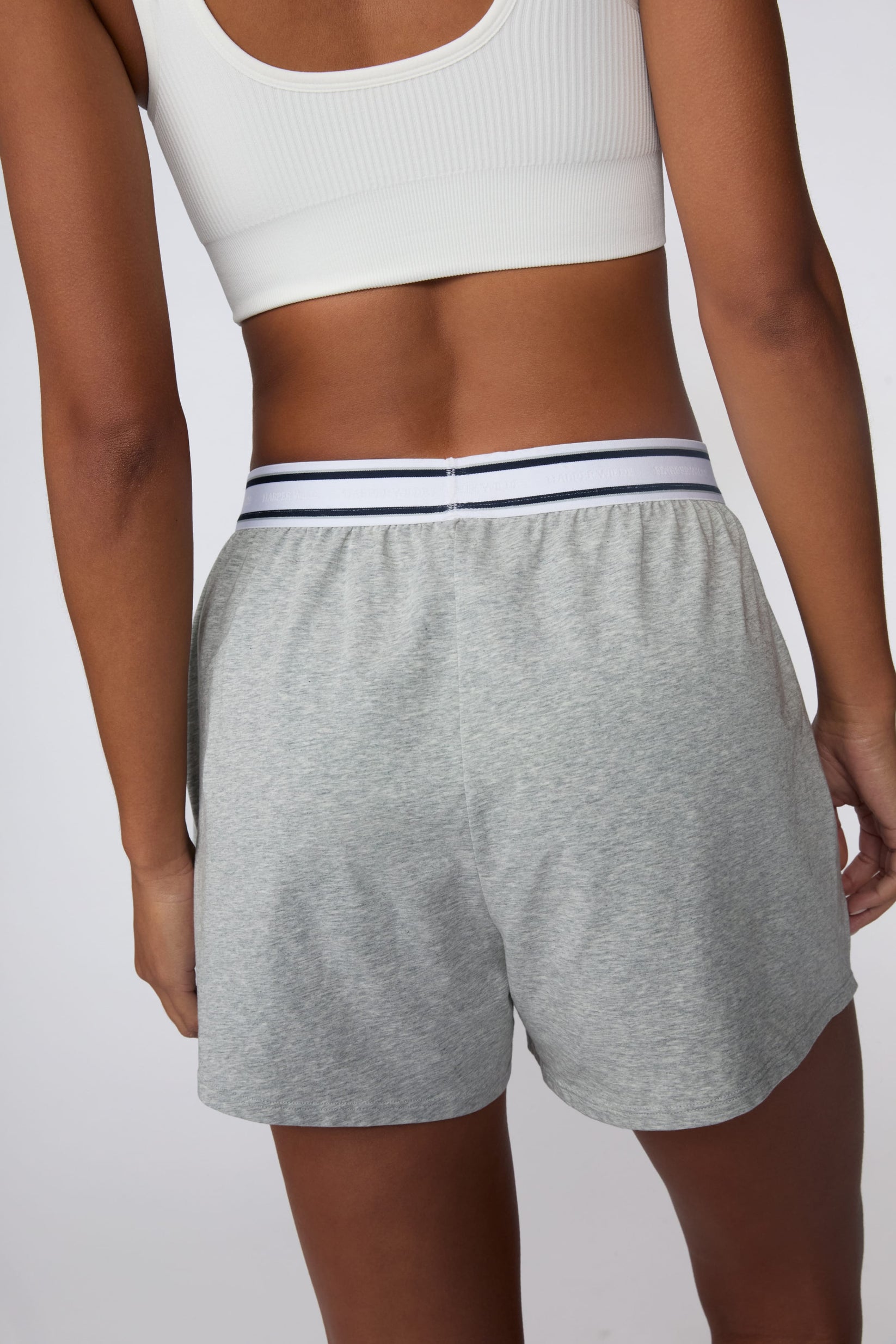 On a light gray studio backdrop, Amber wears the Cloud Cotton Pajama Short in Heather Grey with a white ribbed bralette. The back view highlights the relaxed fit, soft cotton fabric, and striped elastic waistband.