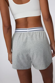 On a light gray studio backdrop, Amber wears the Cloud Cotton Pajama Short in Heather Grey with a white ribbed bralette. The back view highlights the relaxed fit, soft cotton fabric, and striped elastic waistband.