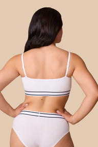 On a neutral beige backdrop, a back view of Emersyn wearing the Cloud Cotton Retro Bandeau Bralette in White showcases the straight back design, adjustable straps, and striped elastic underband.