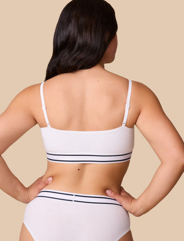 On a neutral beige backdrop, a back view of Emersyn wearing the Cloud Cotton Retro Bandeau Bralette in White showcases the straight back design, adjustable straps, and striped elastic underband.