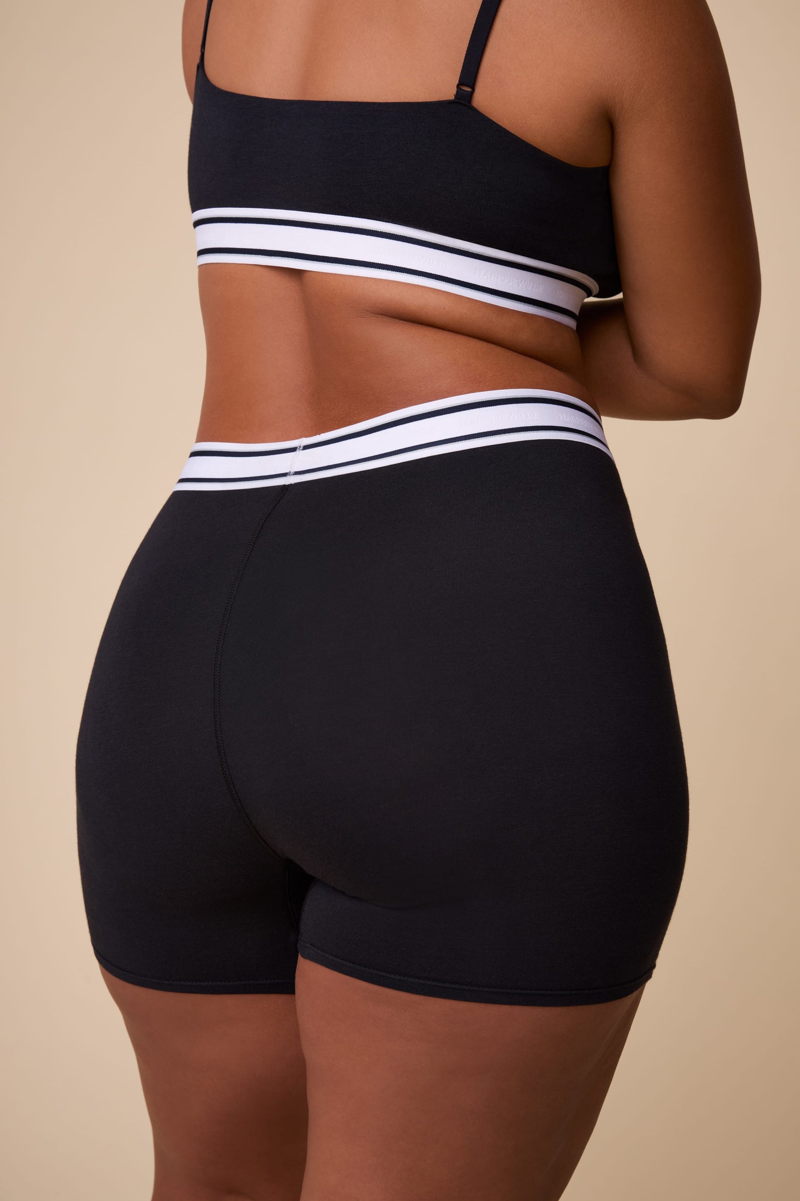 On a neutral studio backdrop, India wears the Cloud Cotton Retro Boxer Brief in Black. The back view highlights the full-coverage fit, smooth cotton modal fabric, and retro-inspired logo waistband with contrasting stripes.