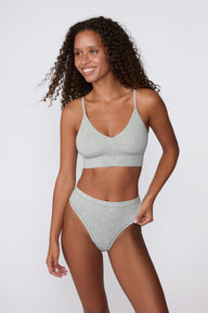 On a light gray studio backdrop, Courtney wears the Cloud Cotton Triangle Bralette in Heather Grey with matching underwear. She smiles while standing at an angle, highlighting the soft cotton fabric, deep V-neckline, and thin adjustable straps.