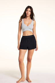On a light studio backdrop, Laura wears the Cloud Cotton Plunge Bralette in Heather Grey with the Cloud Cotton Short in Black. The full-body image highlights the deep V-neck, soft heathered fabric, wide comfort band, and relaxed lounge styling.
