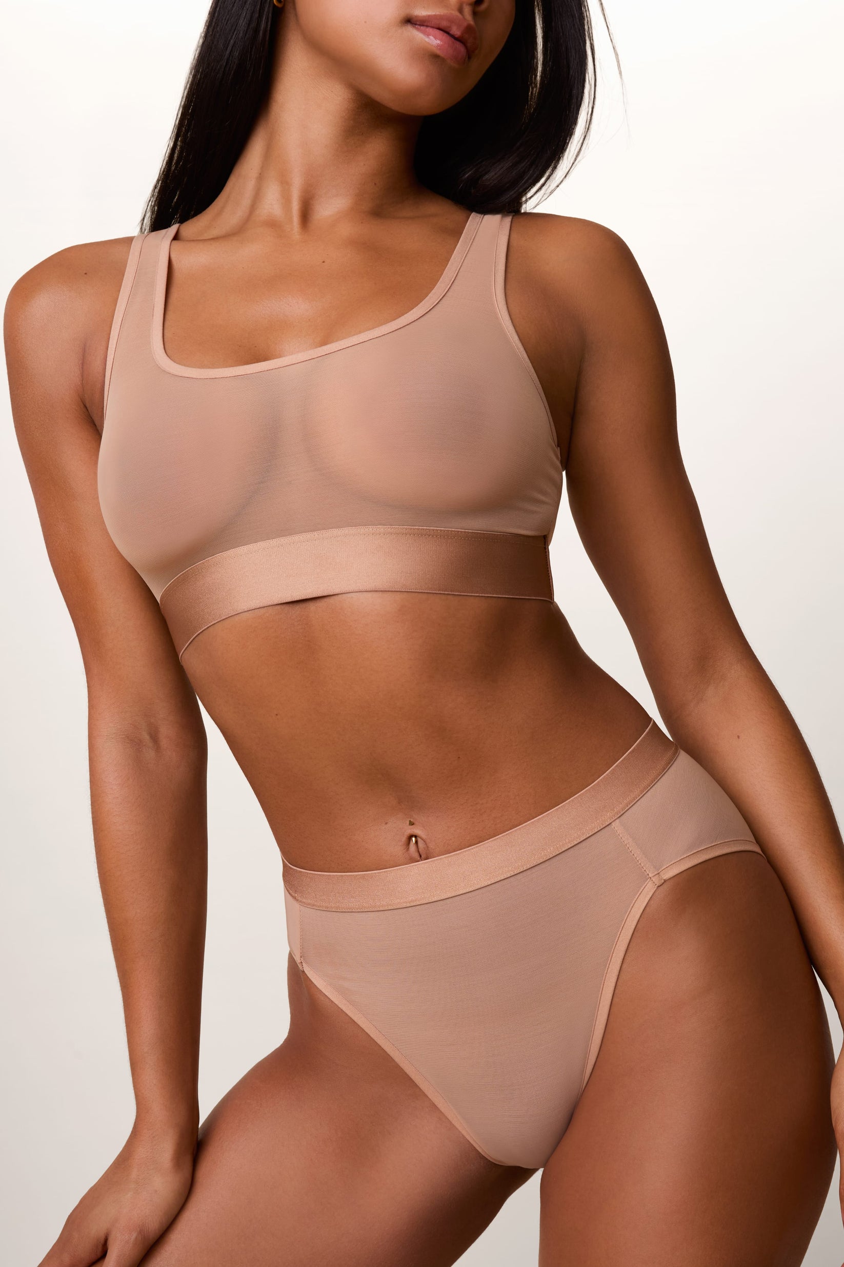 On a light studio backdrop, Ally wears the ComfortMesh™ Scoop Bralette and ComfortMesh™ Highwaist Brief in Taupe. The sheer mesh fabric, scoop neckline, wide underband, and high-rise brief are visible.