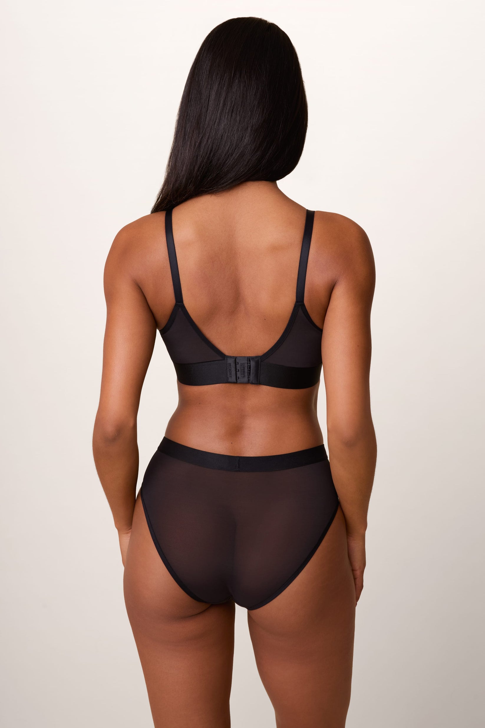 On a light studio backdrop, Ally wears the ComfortMesh™ Triangle Bralette in Black with the matching brief. A back view highlights the adjustable straps, hook-and-eye closure, sheer mesh fabric, and wide comfort band.