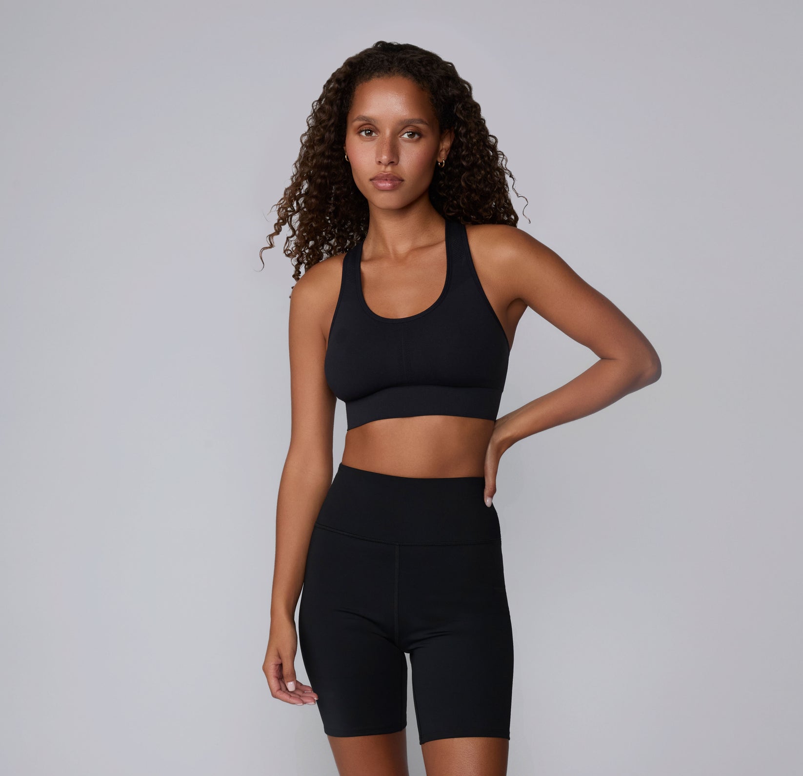 On a neutral studio backdrop, Courtney stands wearing the Seamless Racerback Sports Bra in Black with matching bike shorts. The smooth scoop neckline, wireless support, and clean racerback silhouette are visible from the front.