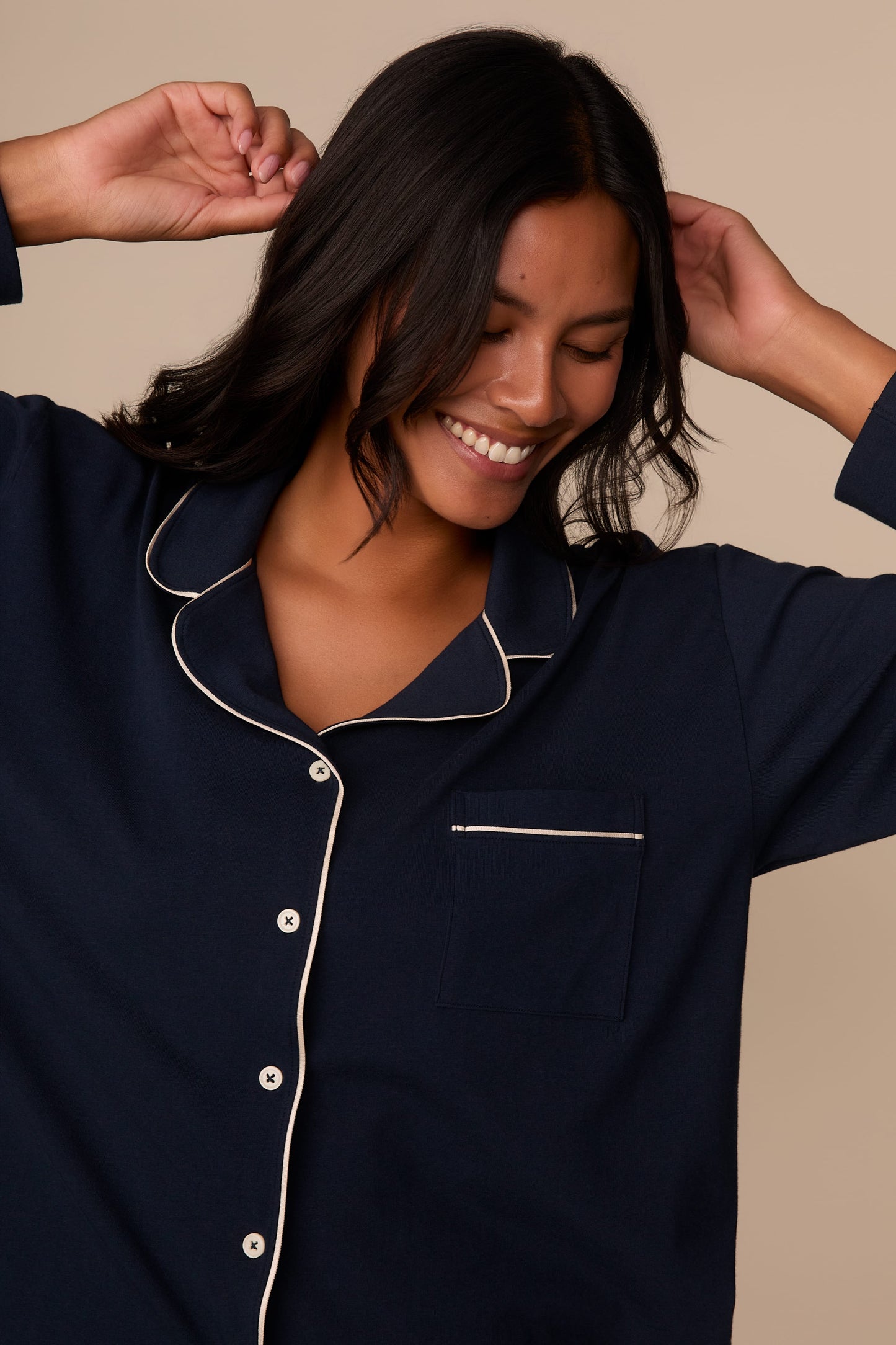 On a neutral studio backdrop, Victoria wears the Perfect Pima Pajama Long Sleeve Shirt in Navy. The close-up view highlights the notched collar, button-front closure, chest pocket, and contrast piping details. Victoria smiles with her hands raised near her hair.