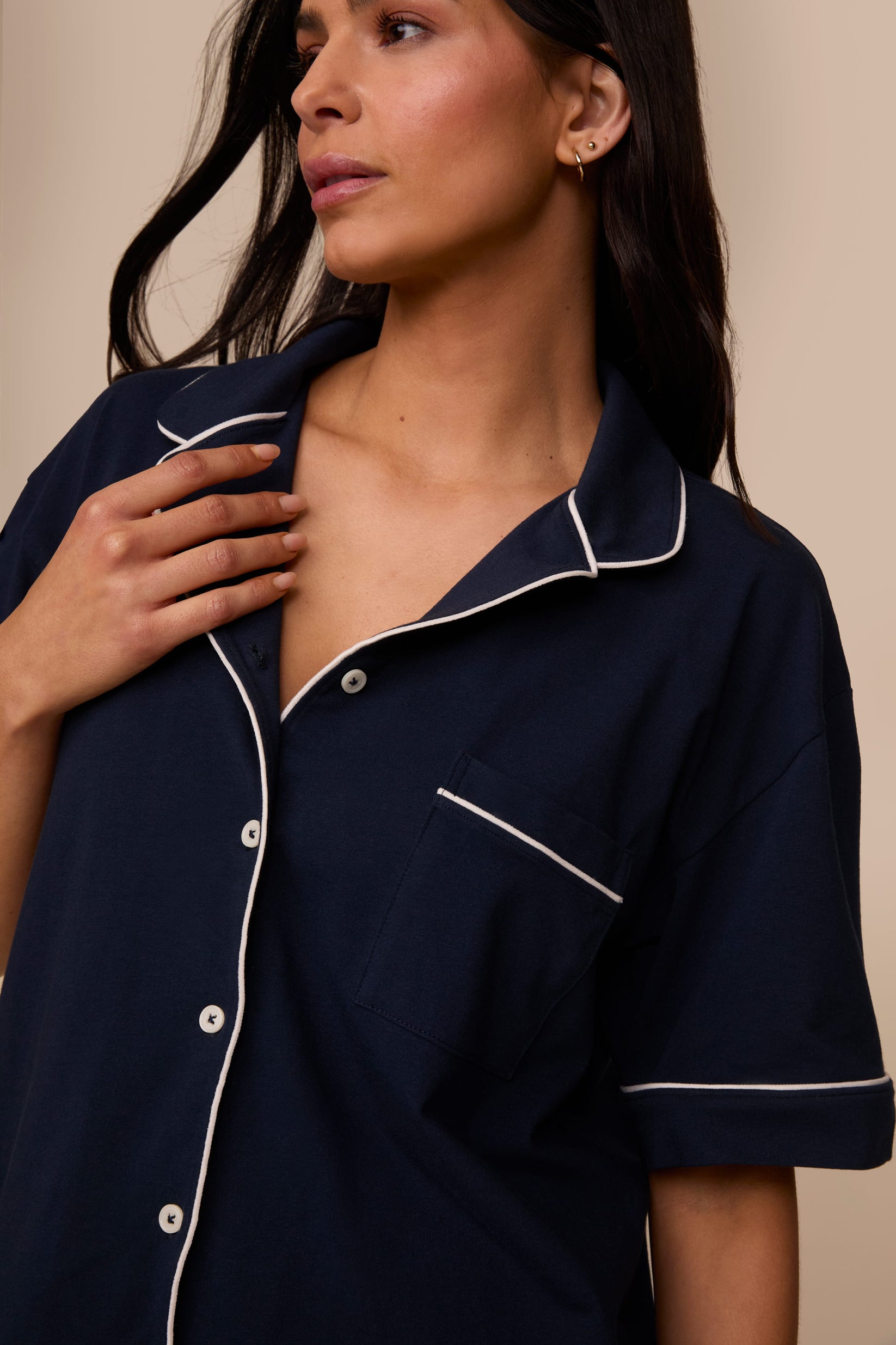 On a neutral studio backdrop, Marcela wears the Perfect Pima Pajama Shirt in Navy. The close-up view highlights the camp collar, button-front closure, chest pocket, and contrast piping along the collar, placket, pocket, and sleeve cuffs.