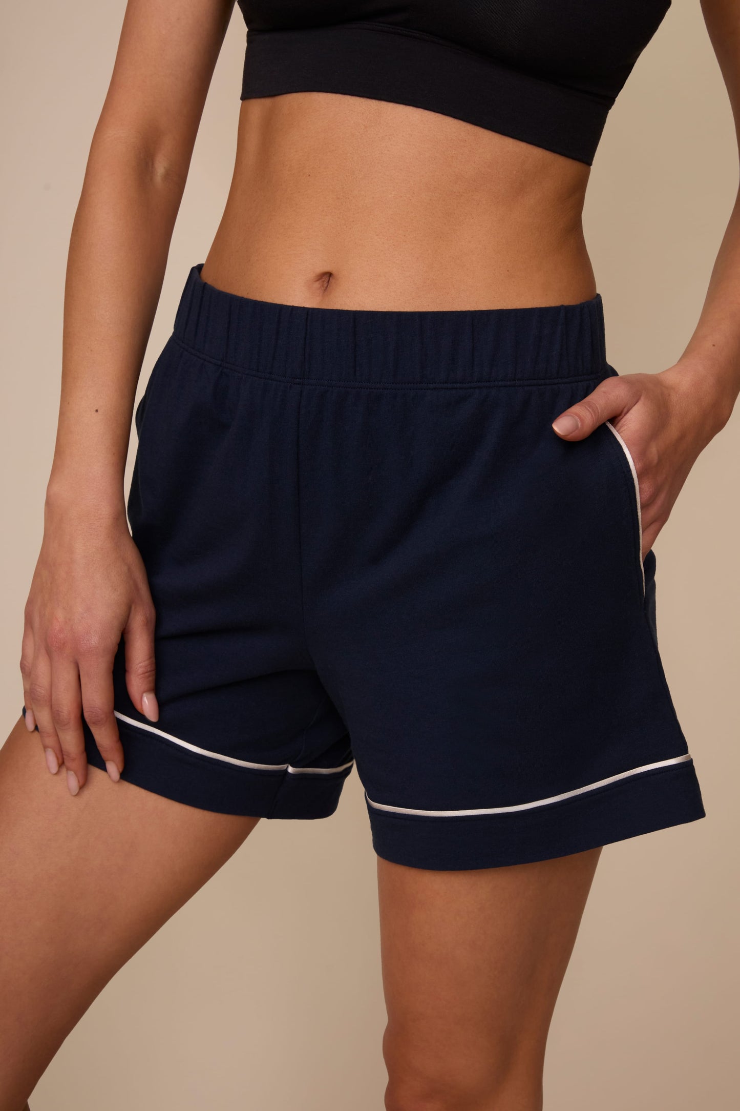On a neutral studio backdrop, Marcela wears the Perfect Pima Pajama Short in Navy. The close-up view highlights the soft elastic waistband, side pocket opening with contrast piping, and contrast trim along the cuffed hem.