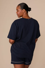 On a warm beige studio backdrop, Zarria wears the Perfect Pima Pajama T-Shirt in a deep navy color with matching pajama shorts. The back view highlights the relaxed fit, short sleeves, and clean, minimalist design.