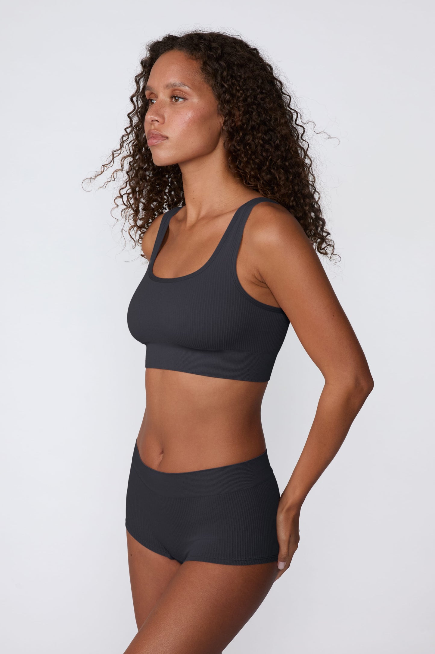 On a light gray studio backdrop, Courtney wears the Ribbed Seamless Scoop Bralette in Charcoal with a matching ribbed bottom. A side profile view highlights the scoop neckline, wide straps, ribbed texture, and supportive underband.