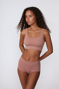 On a light gray studio backdrop, Courtney wears the Ribbed Seamless Scoop Bralette in Plum with a matching ribbed bottom. An angled front view highlights the scoop neckline, wide straps, ribbed texture, and supportive underband.