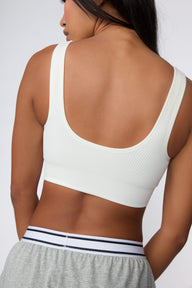 On a light gray studio backdrop, Amber wears the Ribbed Seamless Scoop Bralette in White with gray lounge shorts. A back view highlights the scoop-back design, wide straps, ribbed texture, and supportive underband.